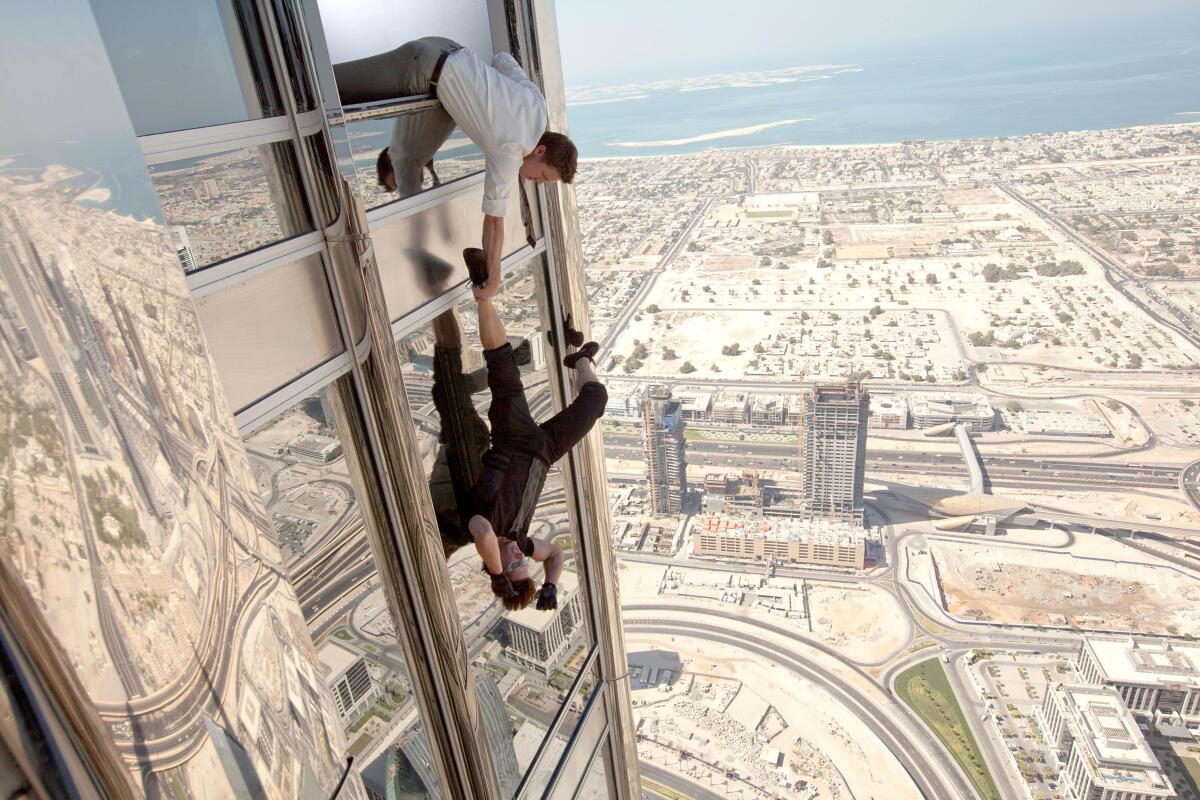 Attempting a rescue, one man is holding the foot of a man who is hanging upside down out of a high-rise, in a movie scene