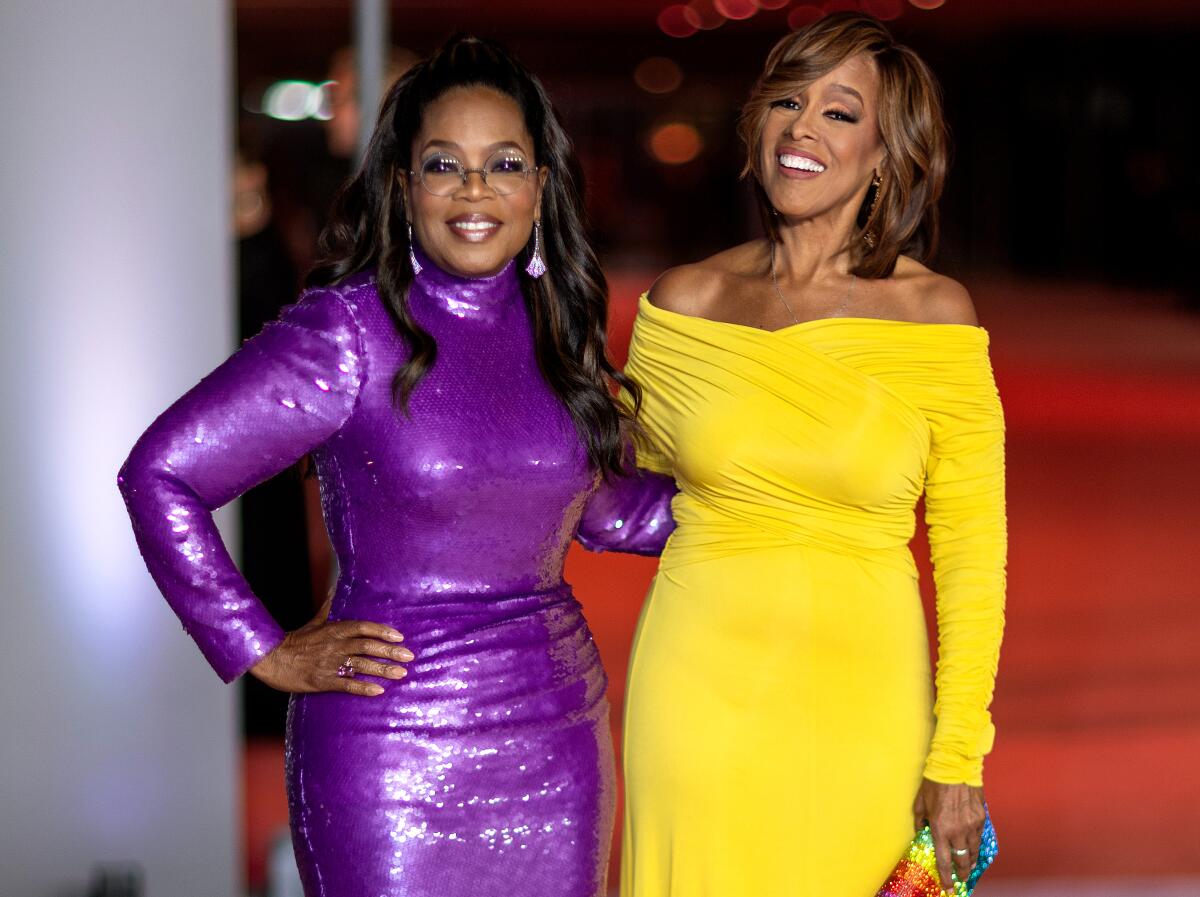 Oprah Winfrey in a shiny purple long-sleeve dress and Gayle King in an off-shoulder yellow long-sleeve dress