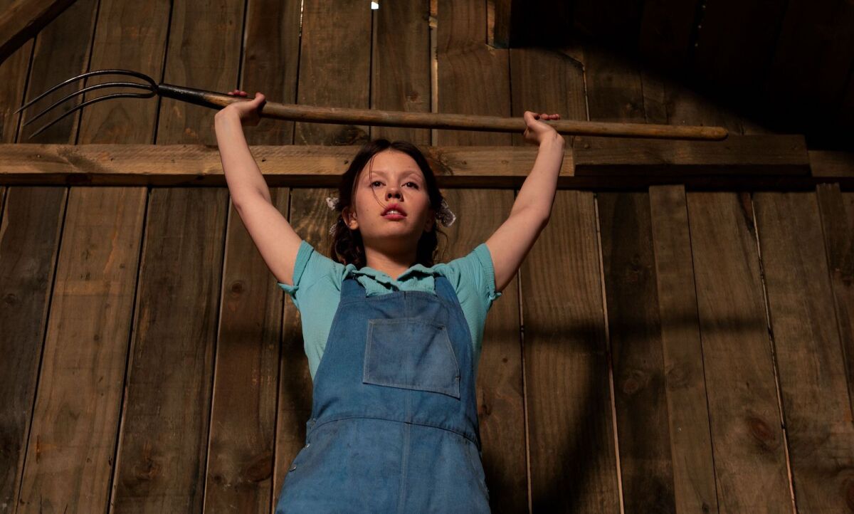 A young woman in overalls raises a pitchfork over her head in the movie "Pearl."