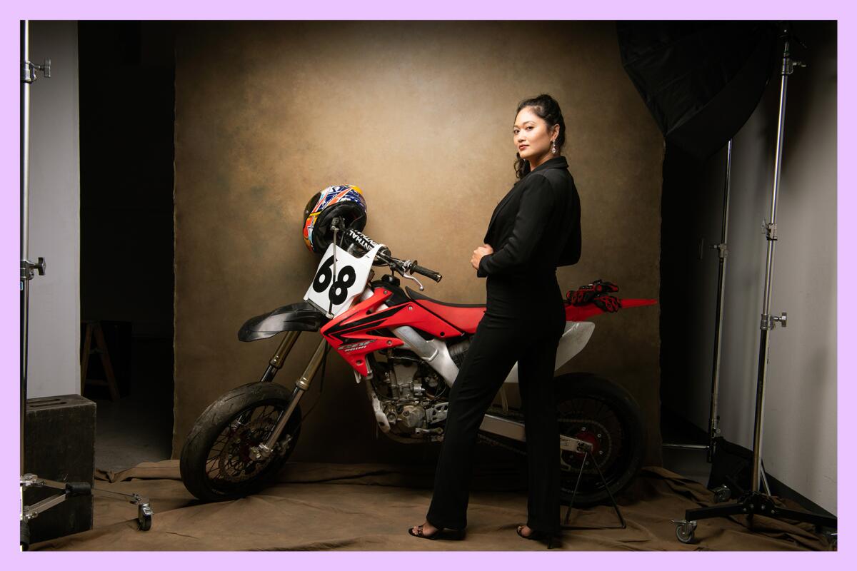A woman dressed in all black poses in a studio with a dirt bike.