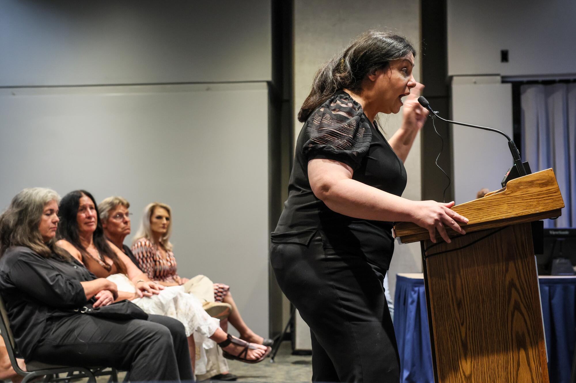 A woman speaks into a microphone at a lectern, with four people seated behind her.