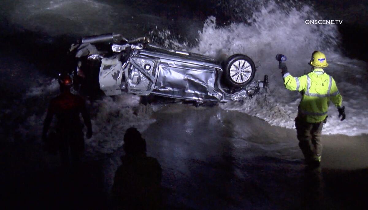 A crashed car is upside down near the ocean as waves crash onto it. A firefighter stands nearby.
