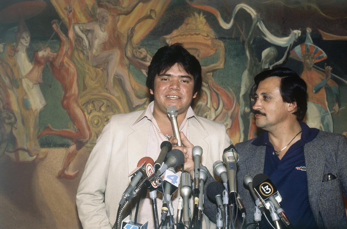 Two men stand in front of a mural, with a tangle of media microphones in front of them