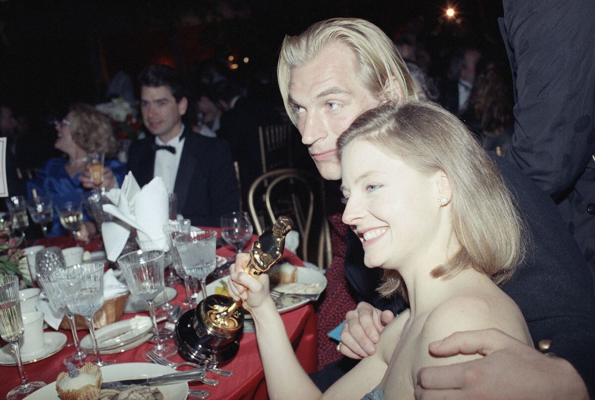 A blond man in a suit holds the shoulders of a blond woman in a strapless dress holding an award.
