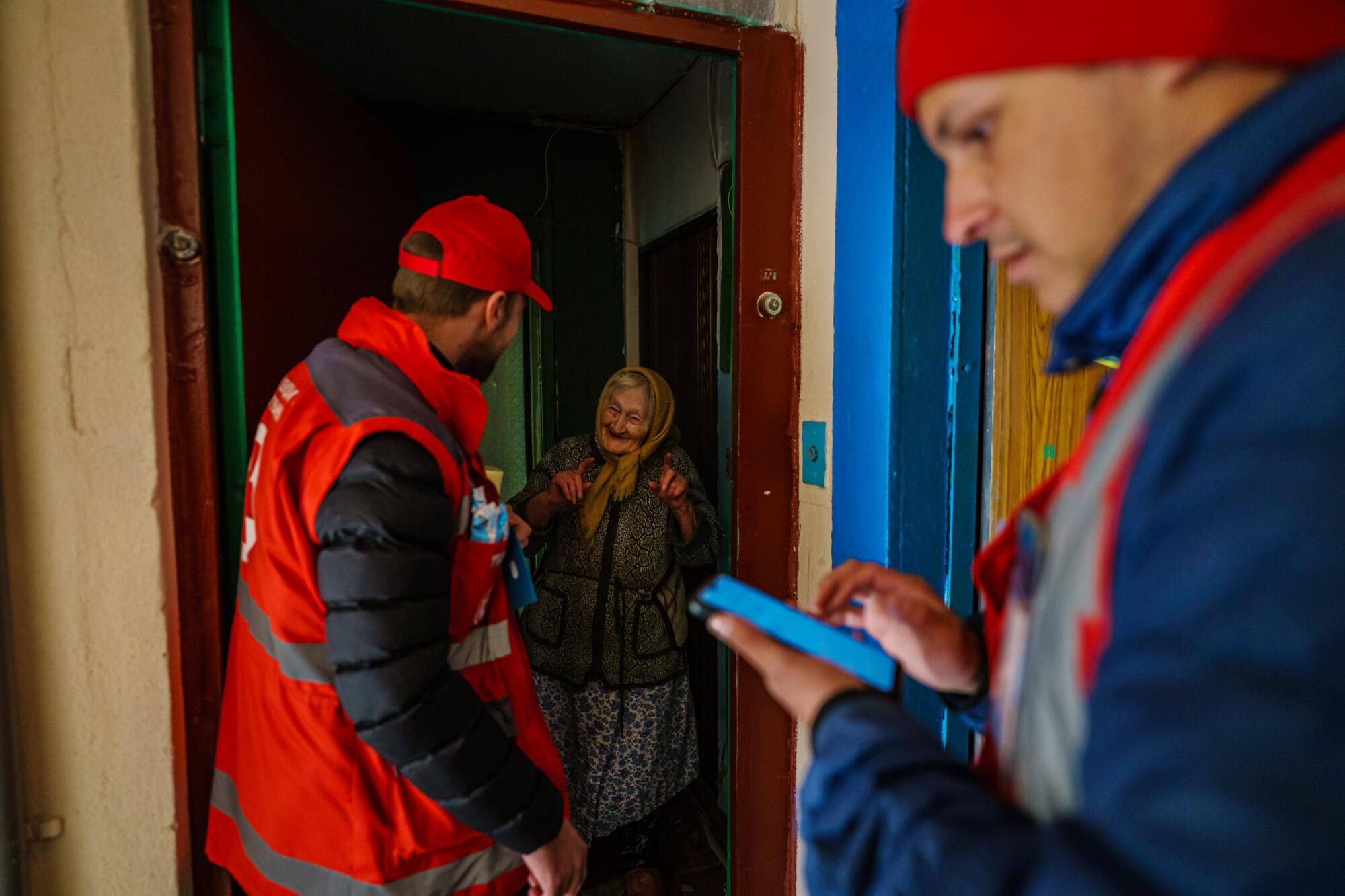 Two male Red Cross workers deliver aid to an elderly female resident in the doorway of her home.