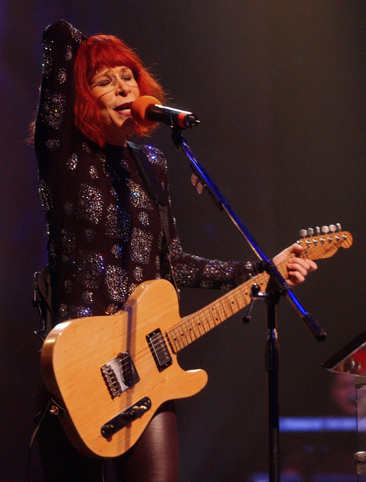 Rita Lee sings into a microphone while playing the guitar