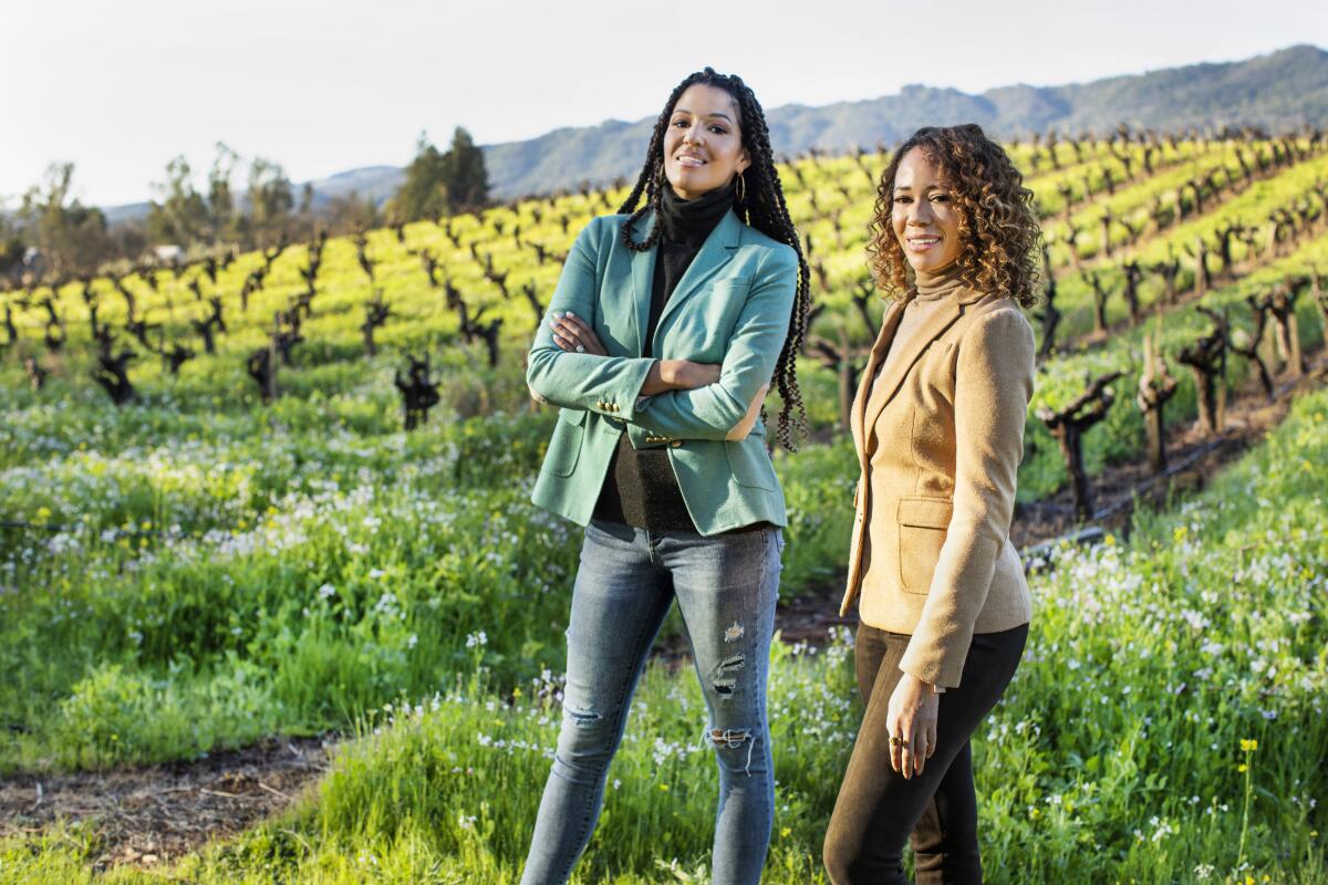 Two women stand and smile in front of a vineyard