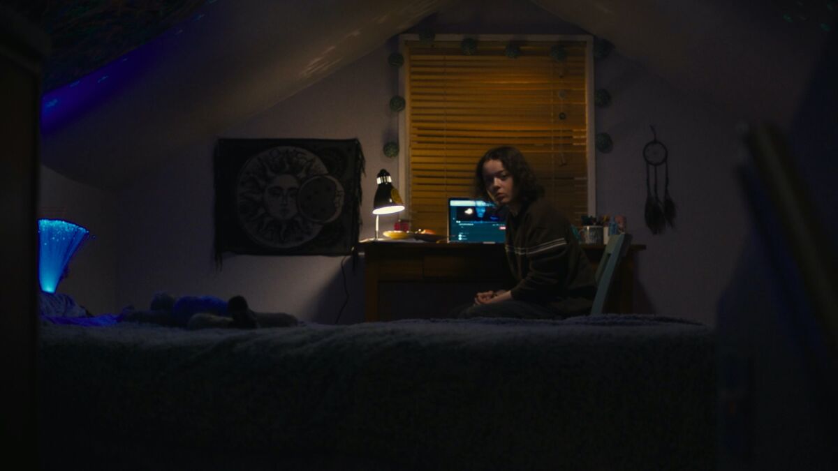A teenage girl sits in her attic bedroom with a computer behind her.