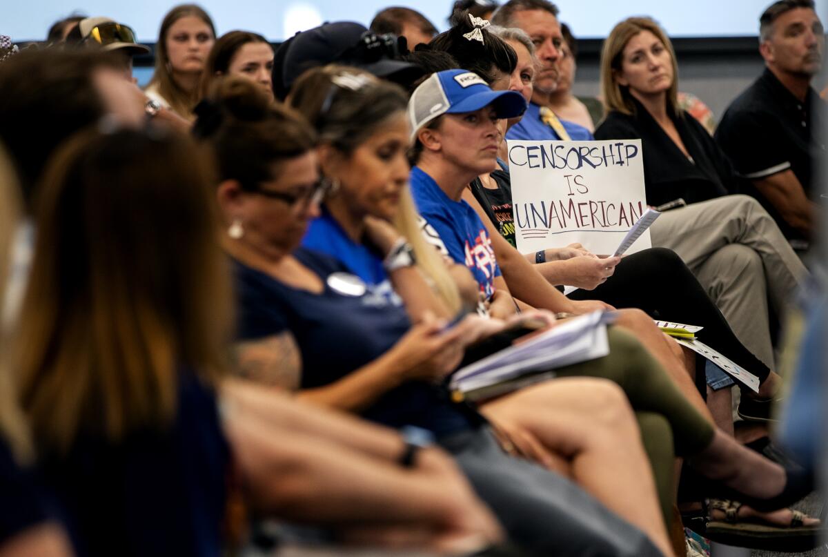 A crowd of people seated at a meeting, one holding a handmade sign reading "CENSORSHIP IS UNAMERICAN."