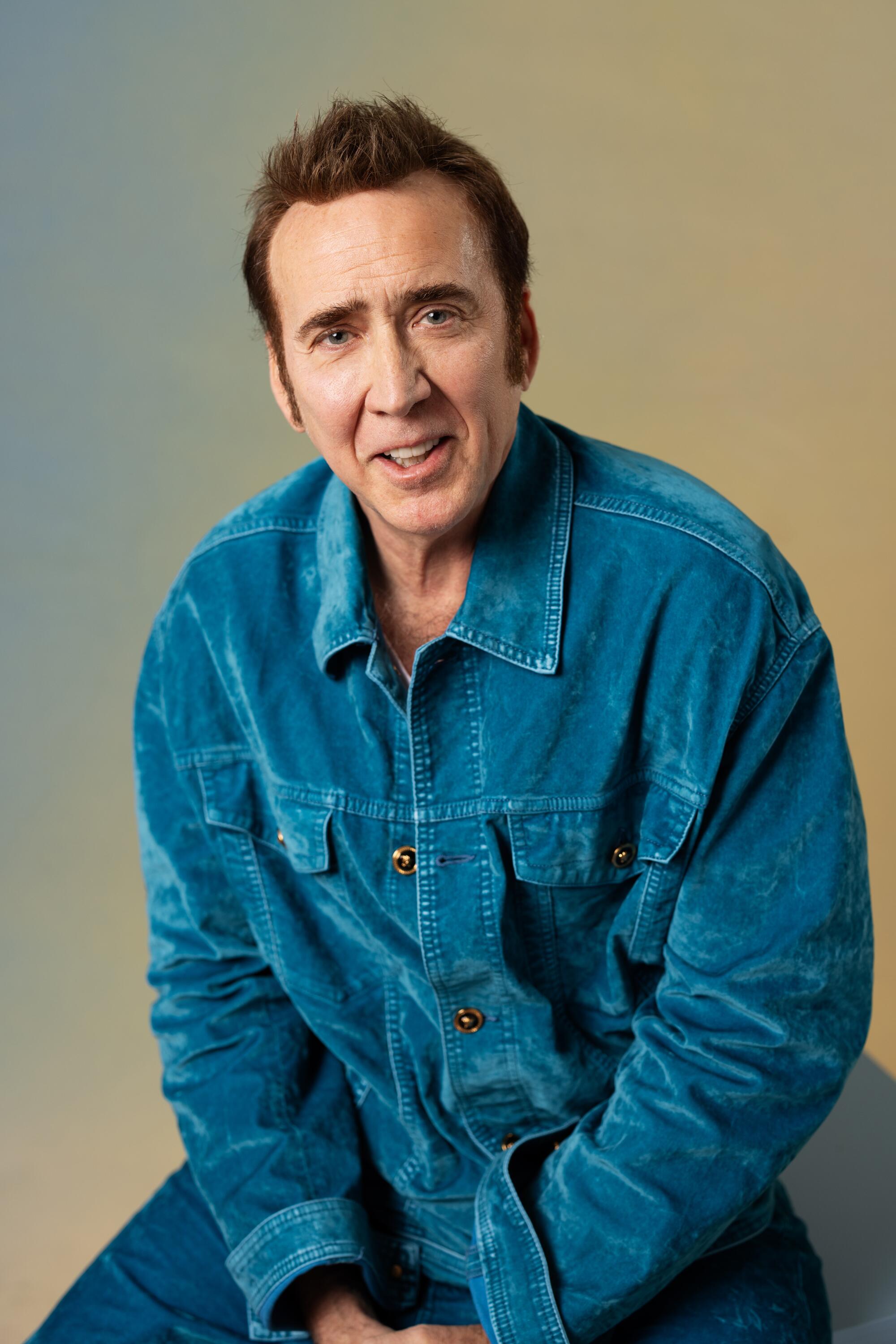 Nicolas Cage sits wearing a blue shirt and smiles.