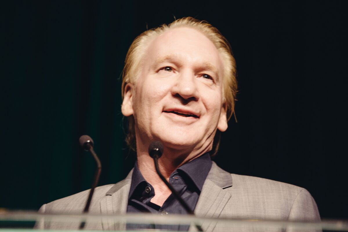 Bill Maher speaks at a podium in front of a black backdrop.