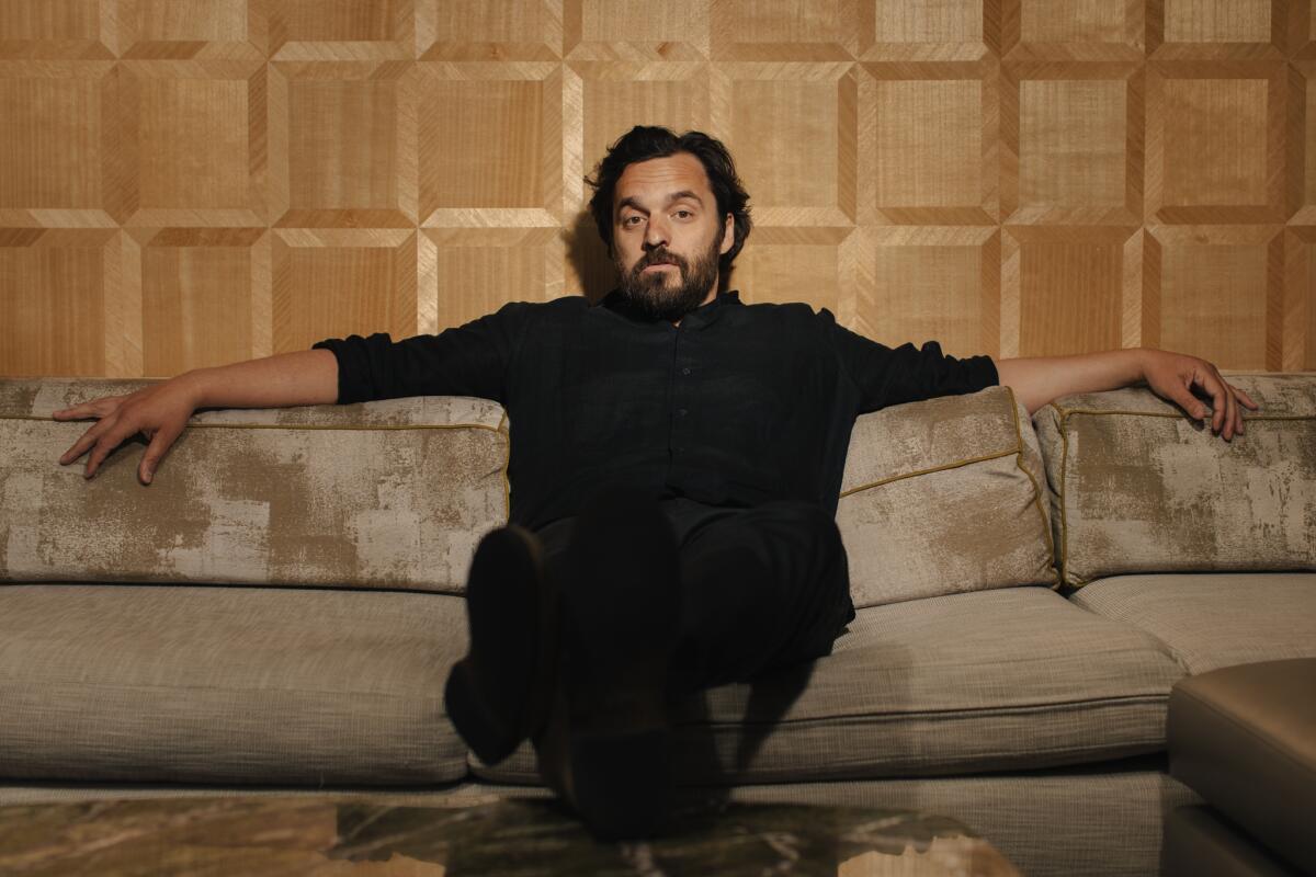 A man wearing black sits on a beige couch with his arms across the top.