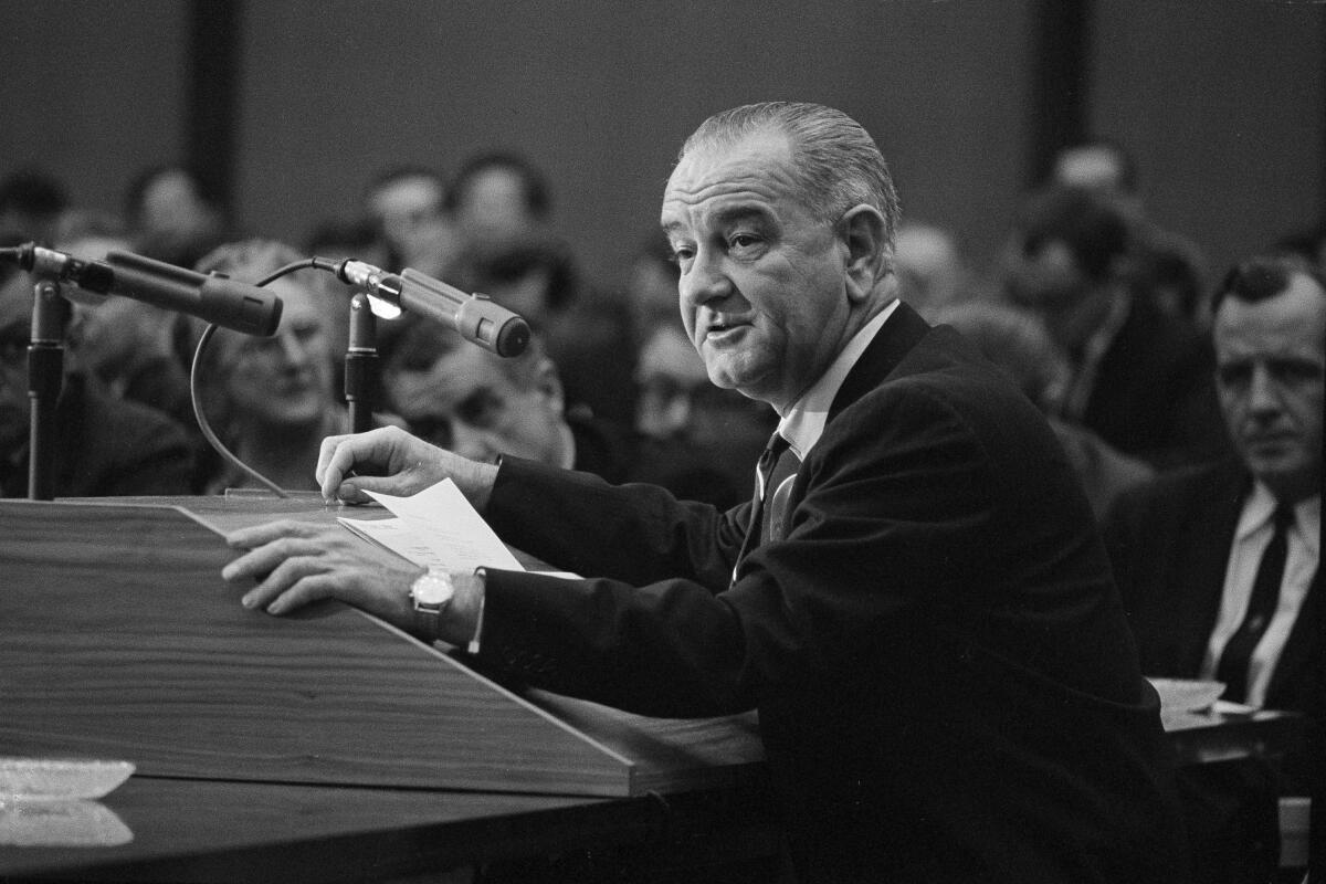 A black and white photo of President Lyndon B. Johnson at a podium with people observing