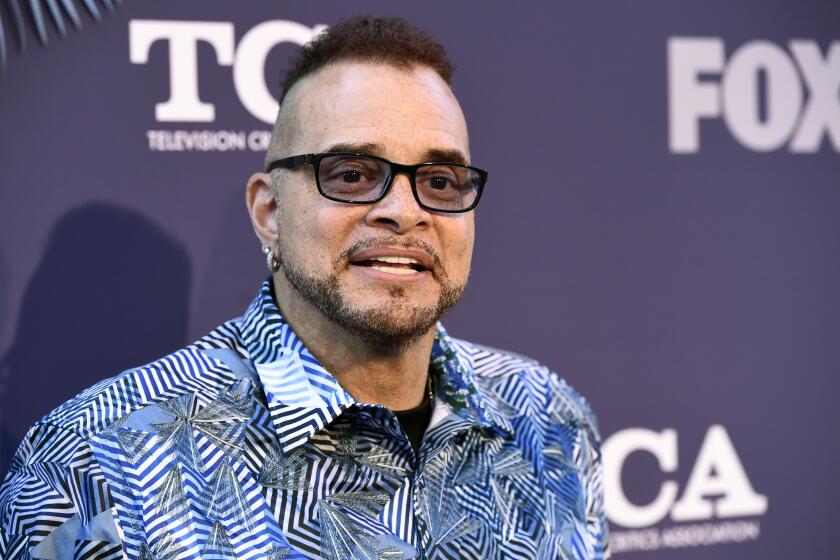 Sinbad with sunglasses in a blue patterned shirt smiling and posing against a dark blue backdrop