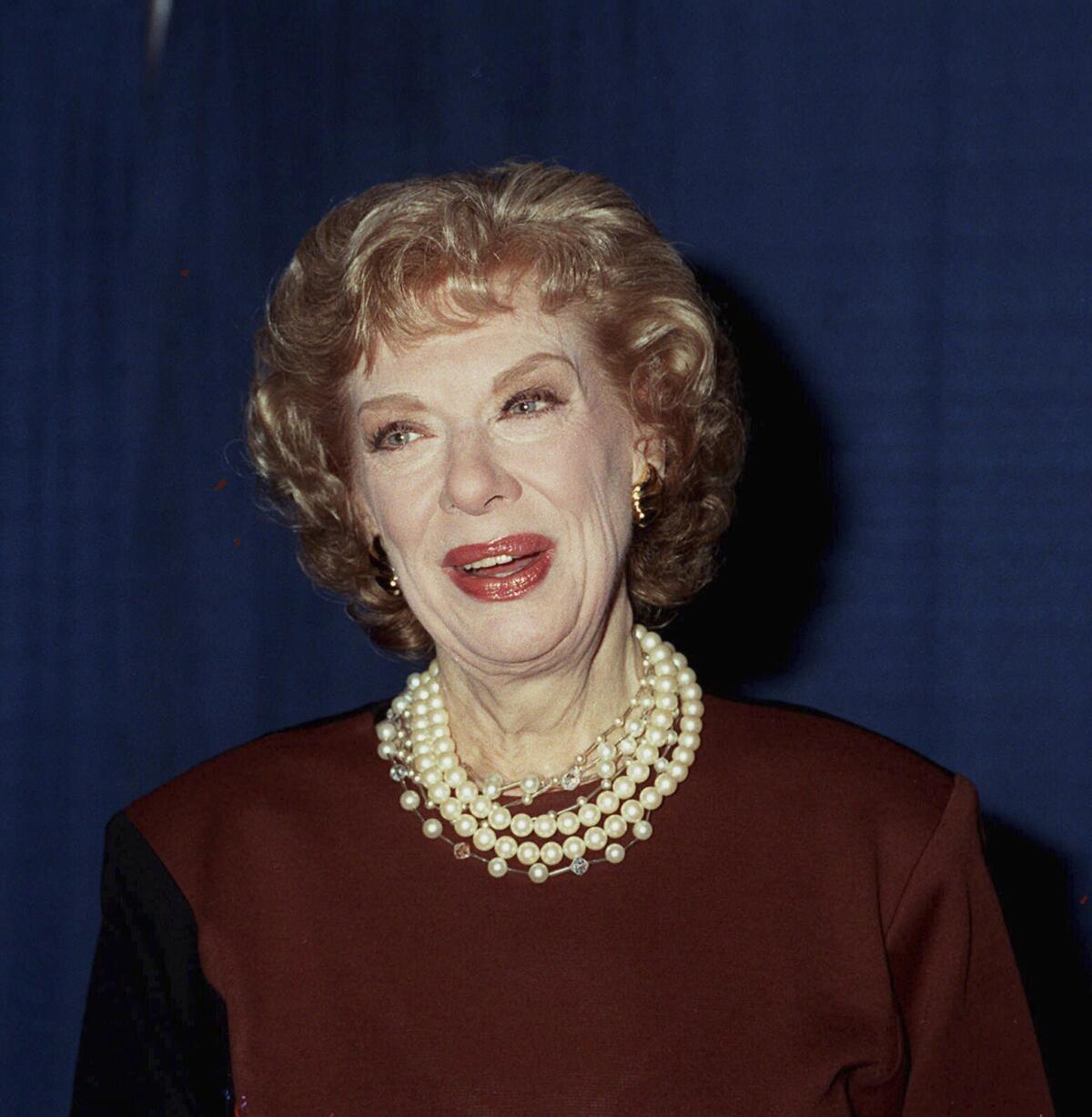 Joyce Randolph wears a burgundy dress with several pearl necklaces as she poses for photos at a red carpet event.