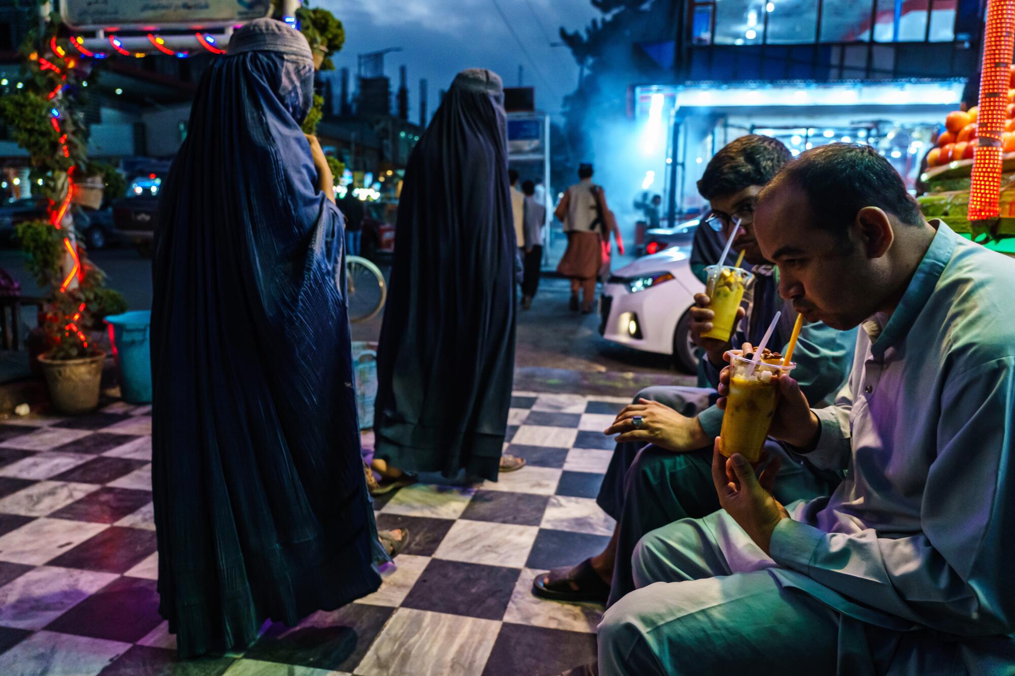 Two men seated at an outdoor area at night as two women in burqas walk by