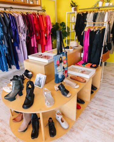 A wooden unit holding shoes in a shop, with colorful clothing hanging on a rack behind