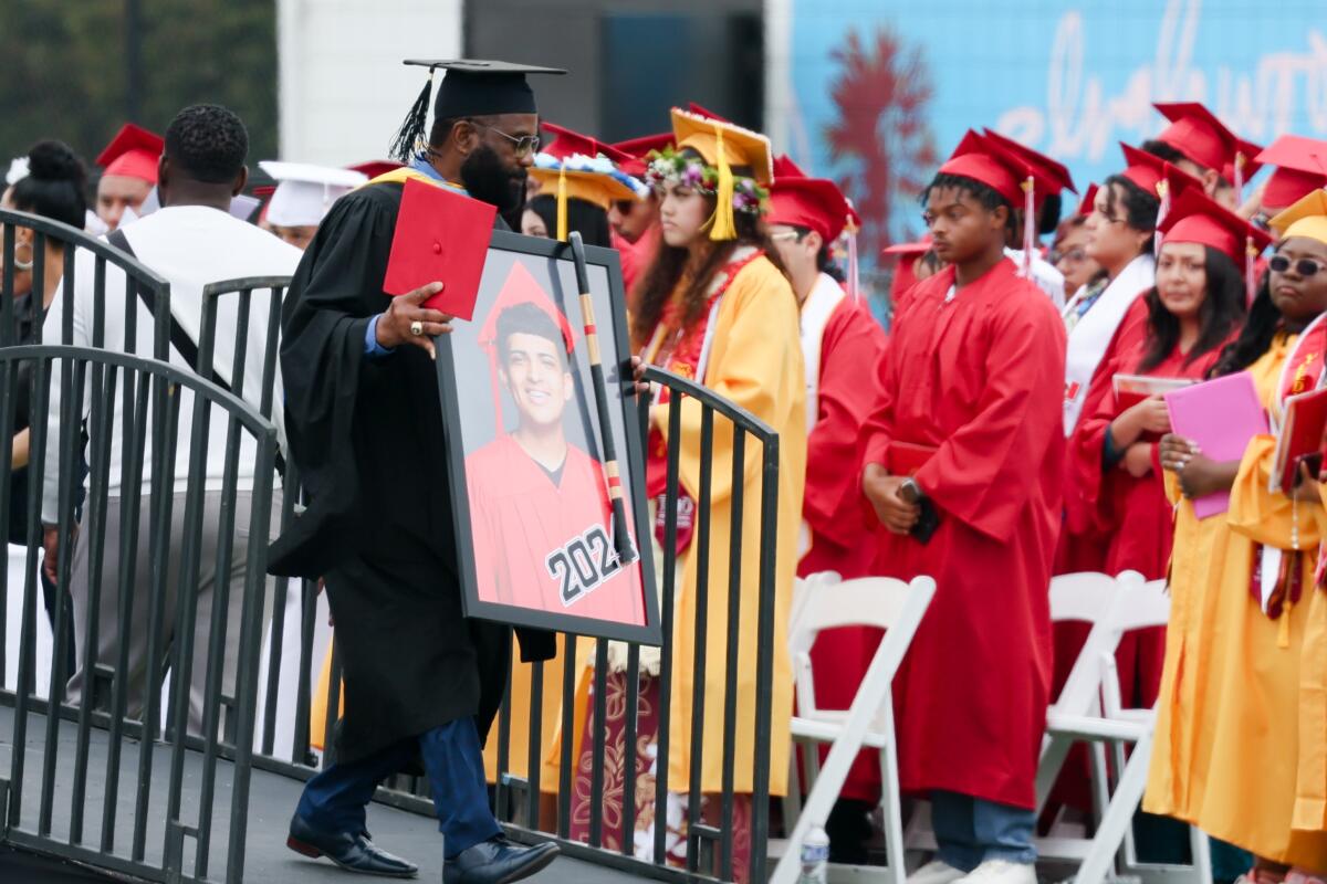 A man in cap and gown holds a large photo of a student in graduation attire.