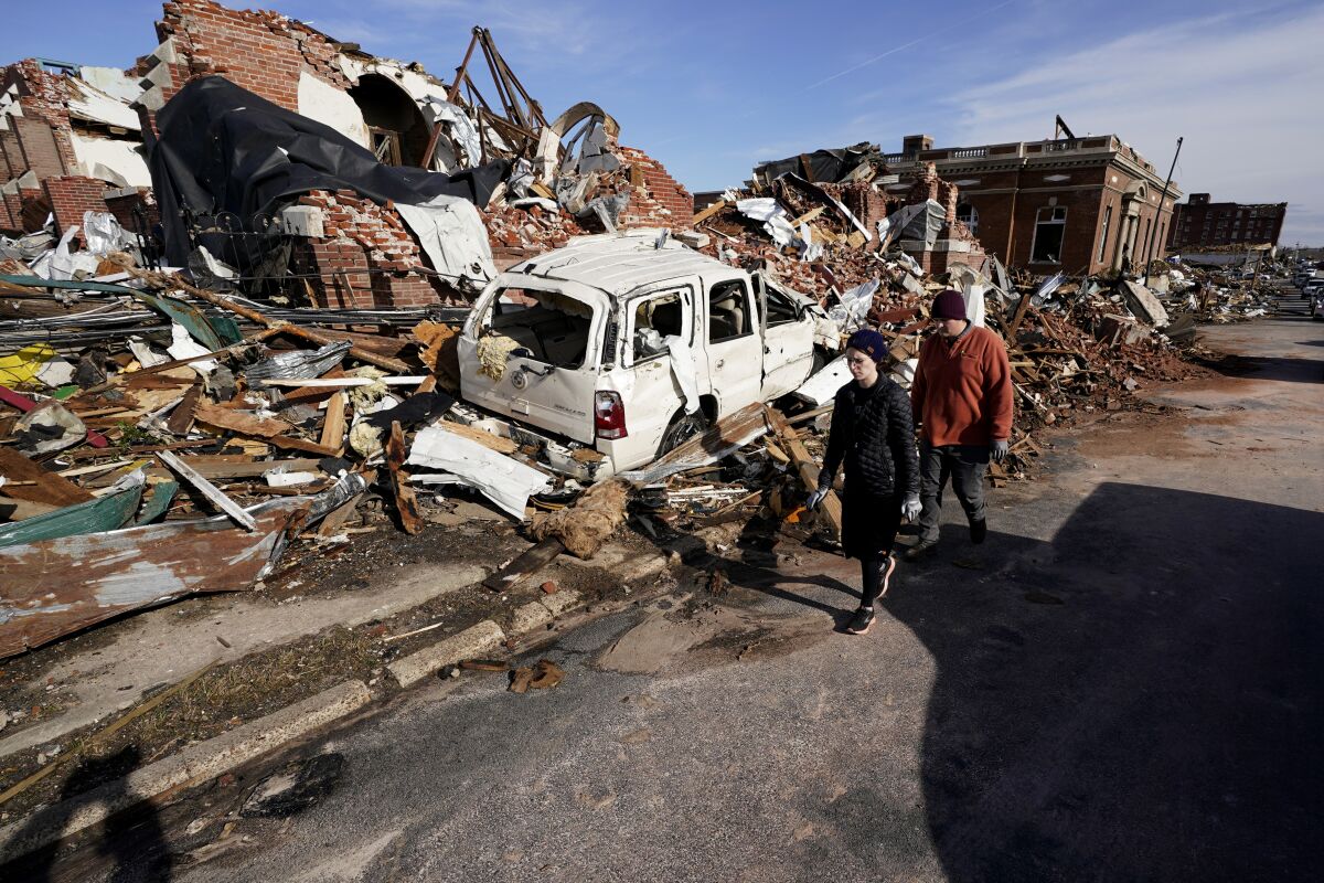 People walk in the street past the remains of a collapsed multistory building and a crushed auto on a heavily damaged street