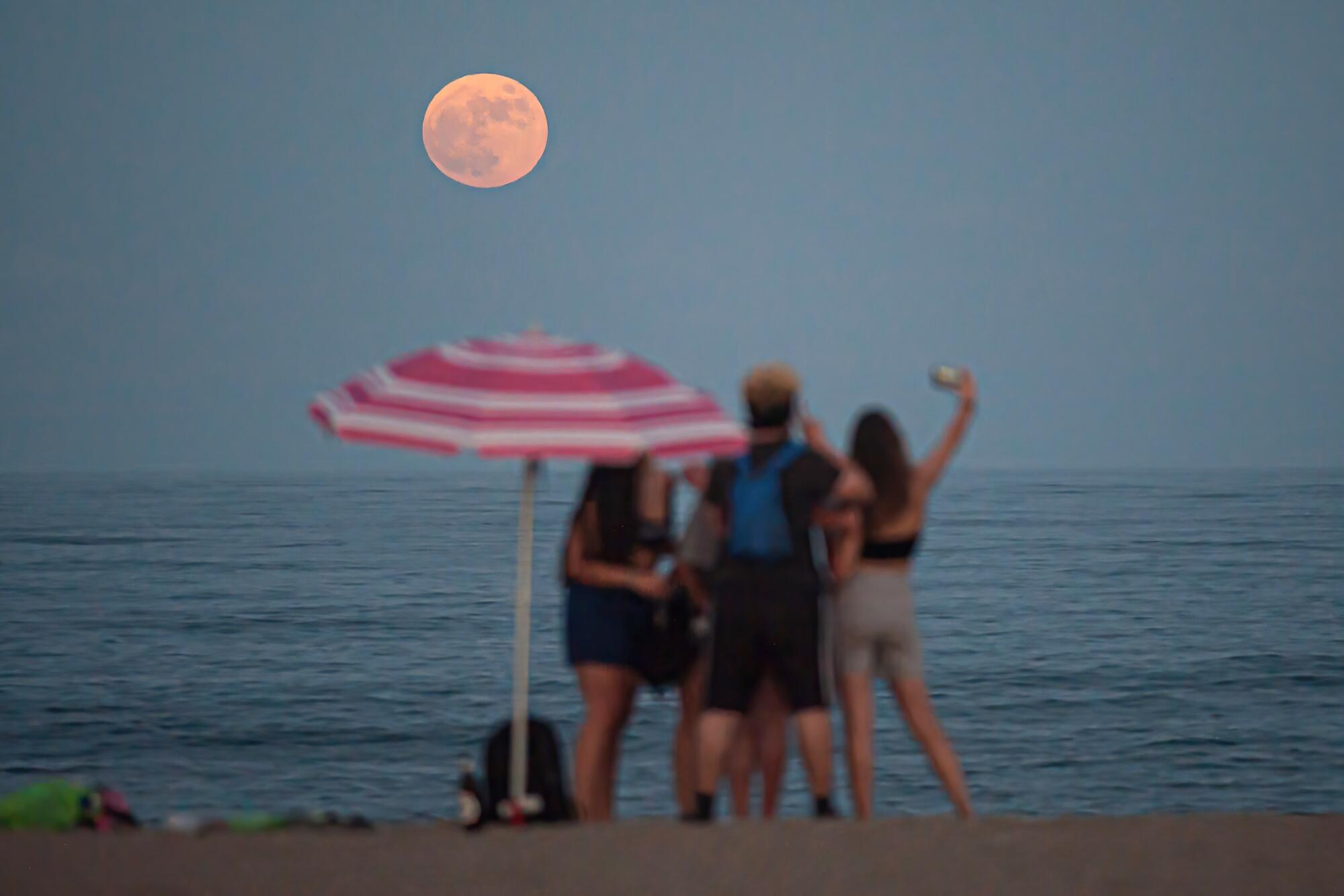 People on a beach watch the full moon rise.