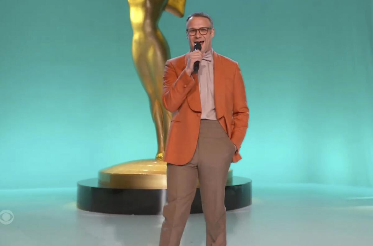 A man with wearing an orange sport coat holds a mic in front of a giant statuette
