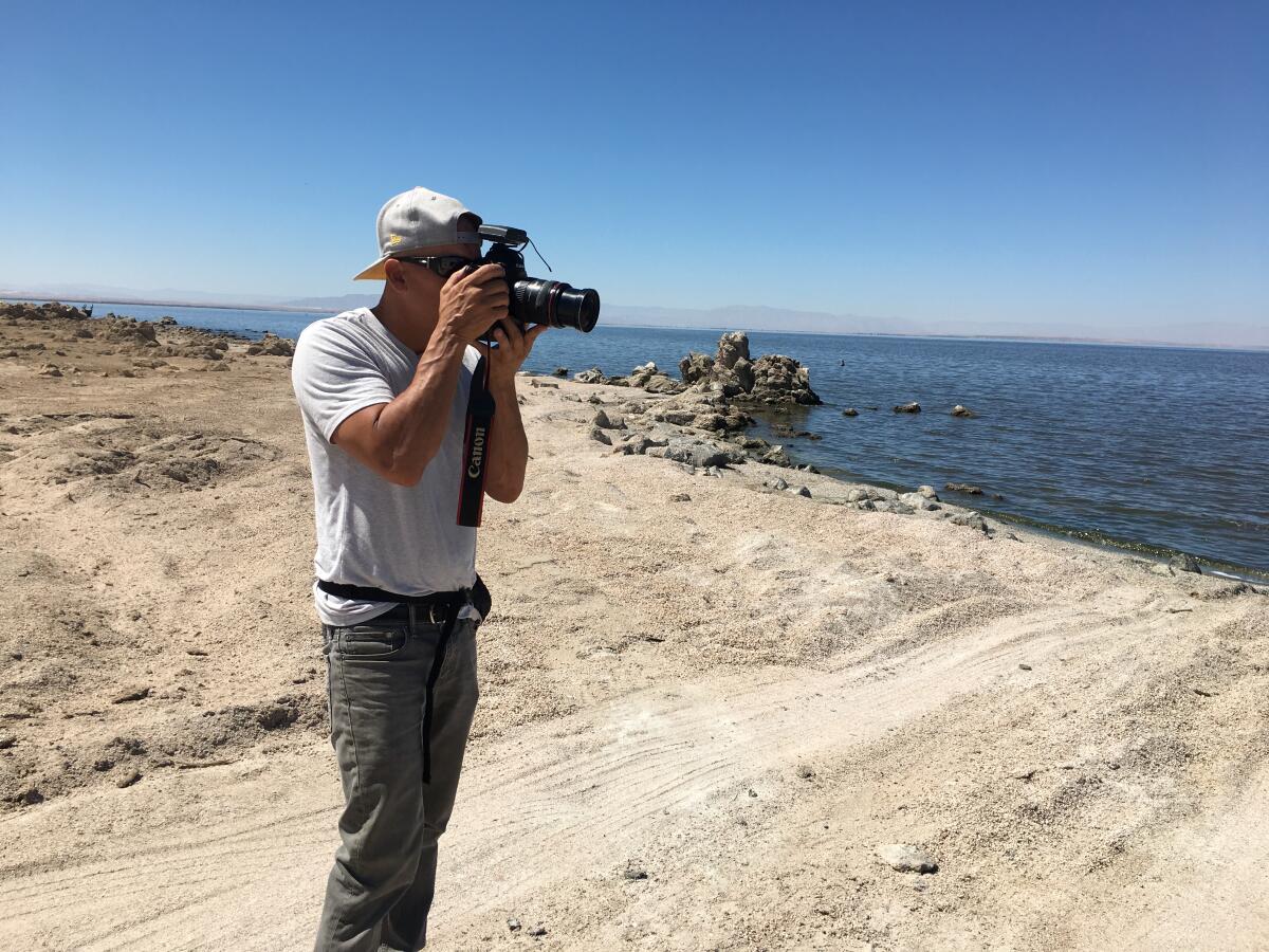 A man in a backward ball cap stands near a body of water and looks through a camera's lens.
