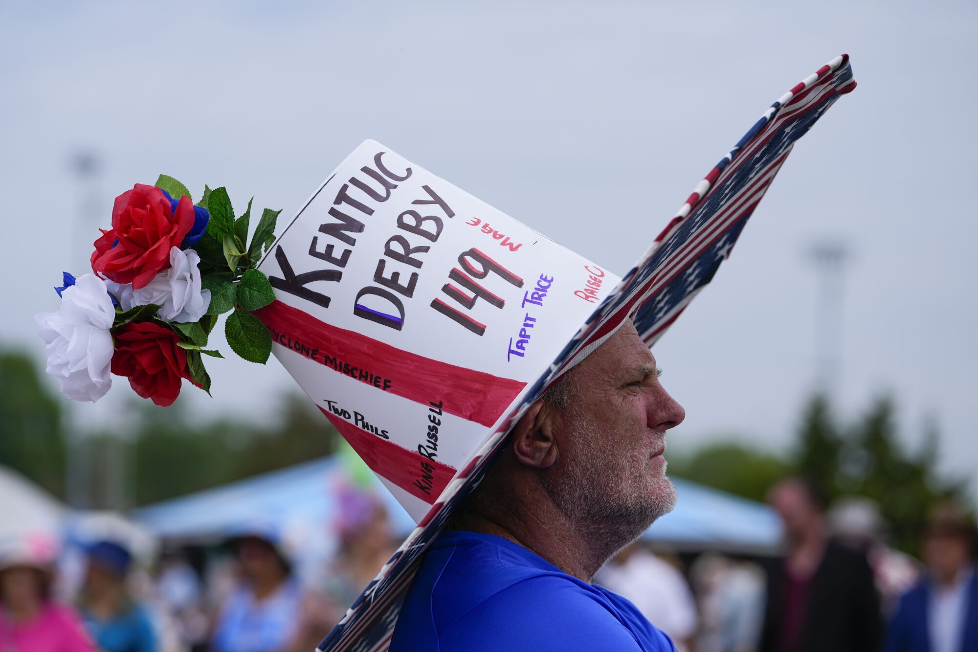 A man wears an oversize top-hat that is white and is covered with the words "Kentucky Derby 149." 