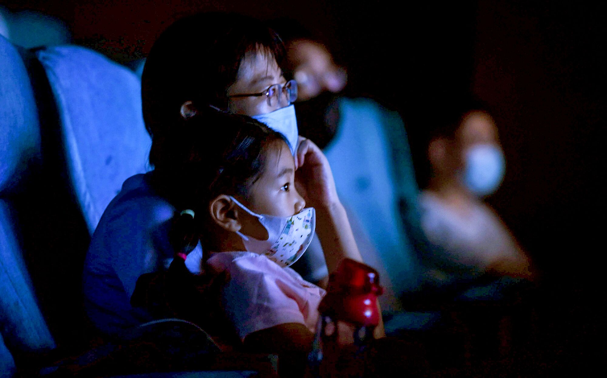 A man and young girl, wearing masks, watch a movie inside a theater.
