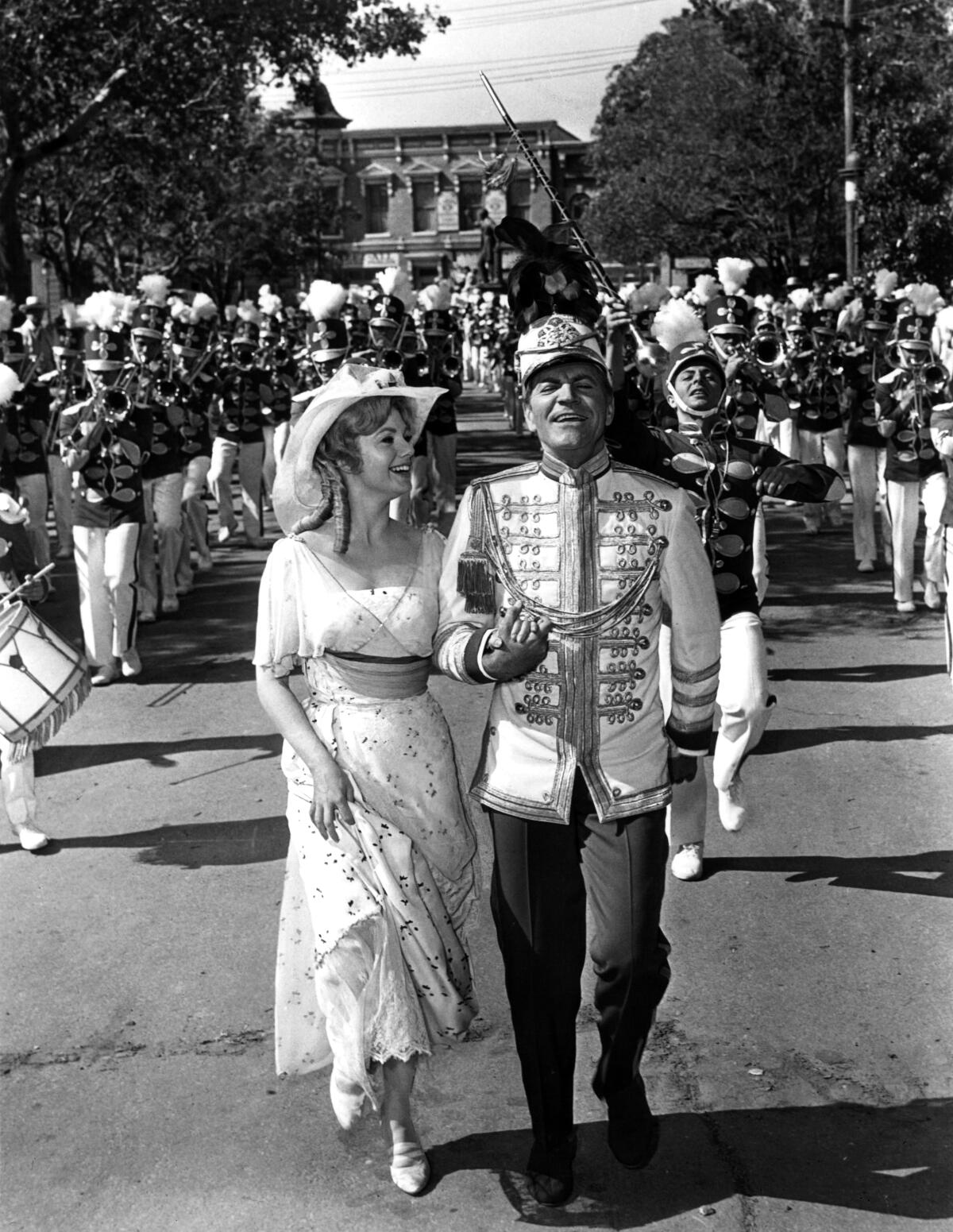 Shirley Jones in period costume and Robert Preston in drum major uniform walk in front of a marching band.