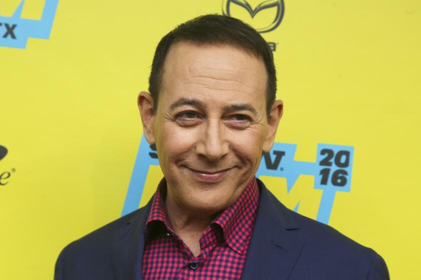 Paul Reubens smiling in a blue suit and checkered red shirt against a yellow backdrop