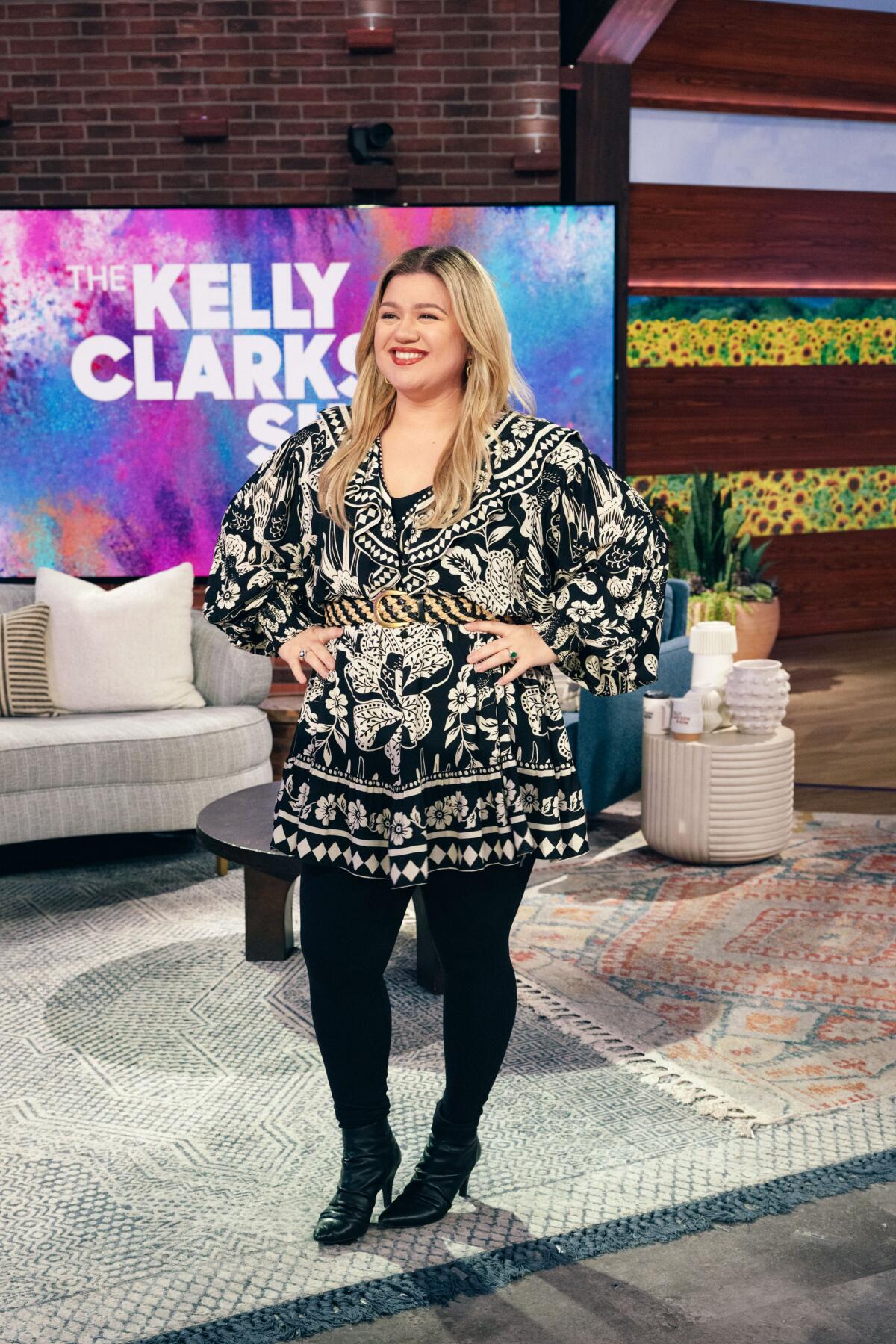 Kelly Clarkson standing on the set of her NBC show with hips on waist, smiling in black and white dress