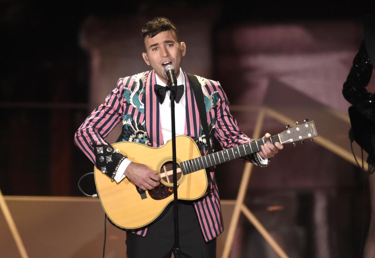 A man in a pink and black striped blazer with a white shirt, black bowtie and black pants plays guitar onstage