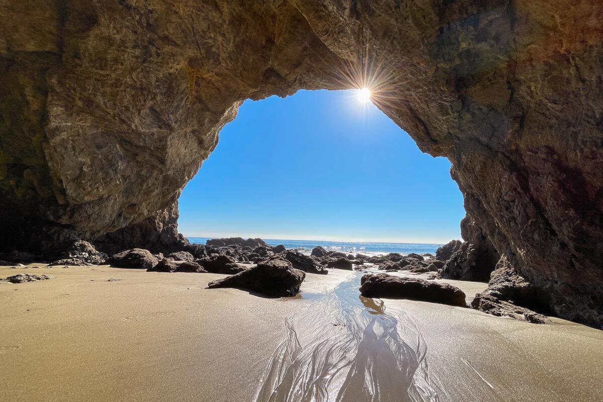 The sun glints through the opening of a cave with a sandy floor.