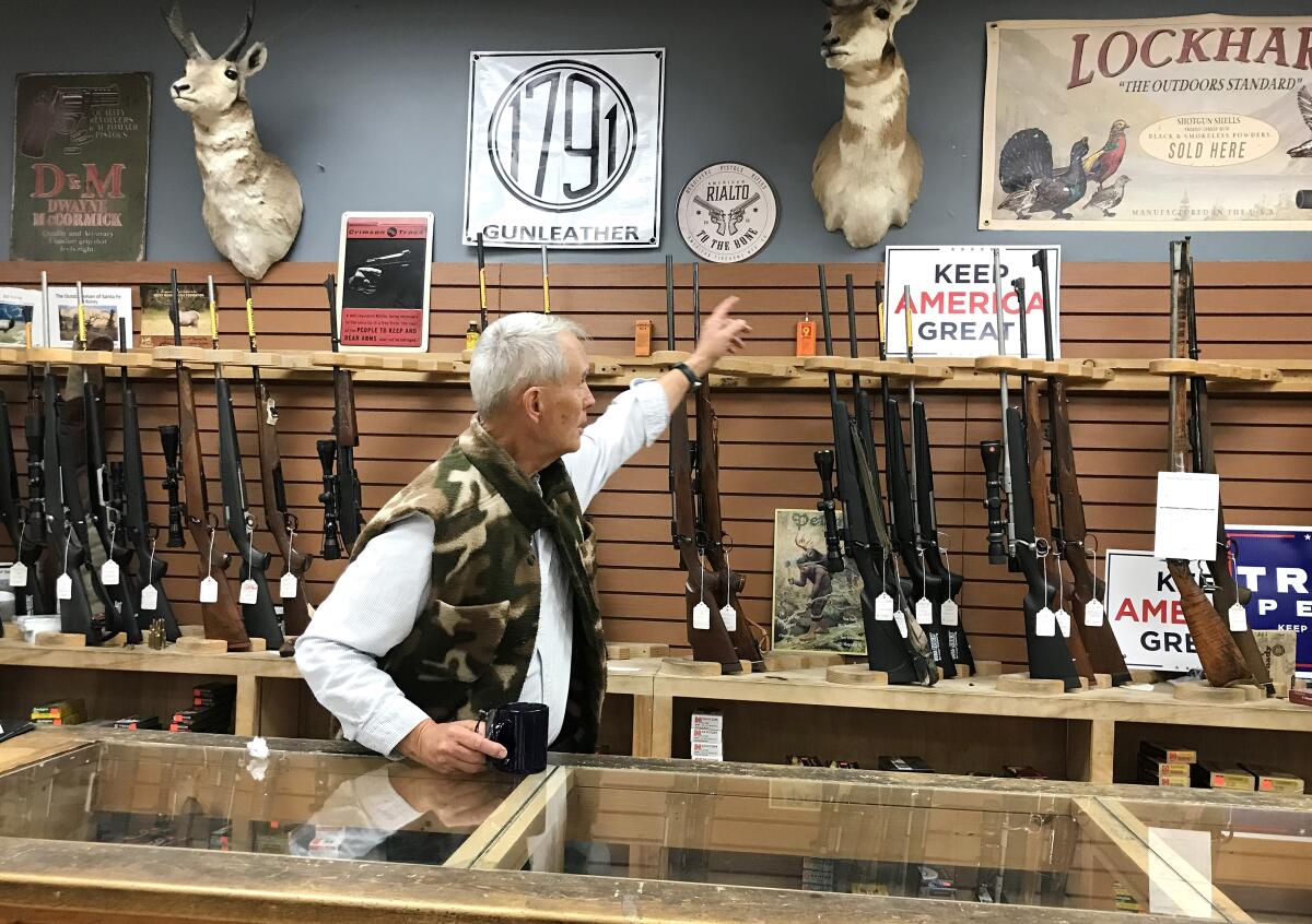 Bill Roney, behind the counter of his gun store in Santa Fe, N.M., reaches toward a wall of rifles and a Trump campaign sign.