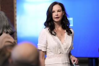 Ashley Judd wears a white dress and stands in front of a blue screen