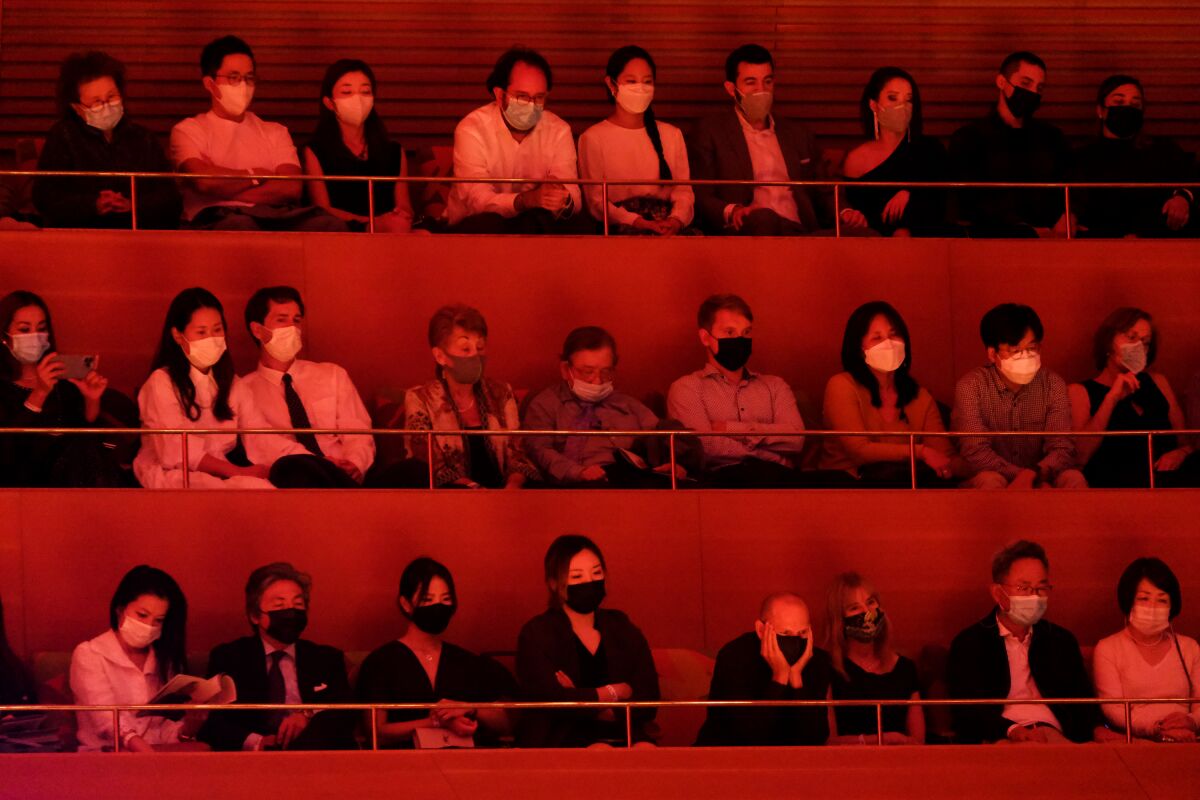 In dim light, people in masks sit in stadium seating in postures of attentiveness.