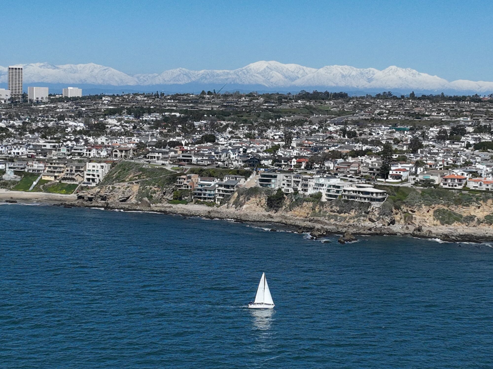 A sailboat cruises off the coast of Corona del Mar with snow-covered mountains in the background.