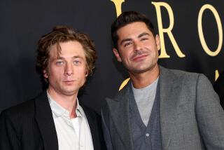 Jeremy Allen White wears a black coat and white shirt and Zac Efron wears a gray coat and shirt as they pose together