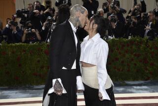 Travis Barker, in a black suit, kisses Kourtney Kardashian, in a white button-up and skirt, with paparrazzi behind them