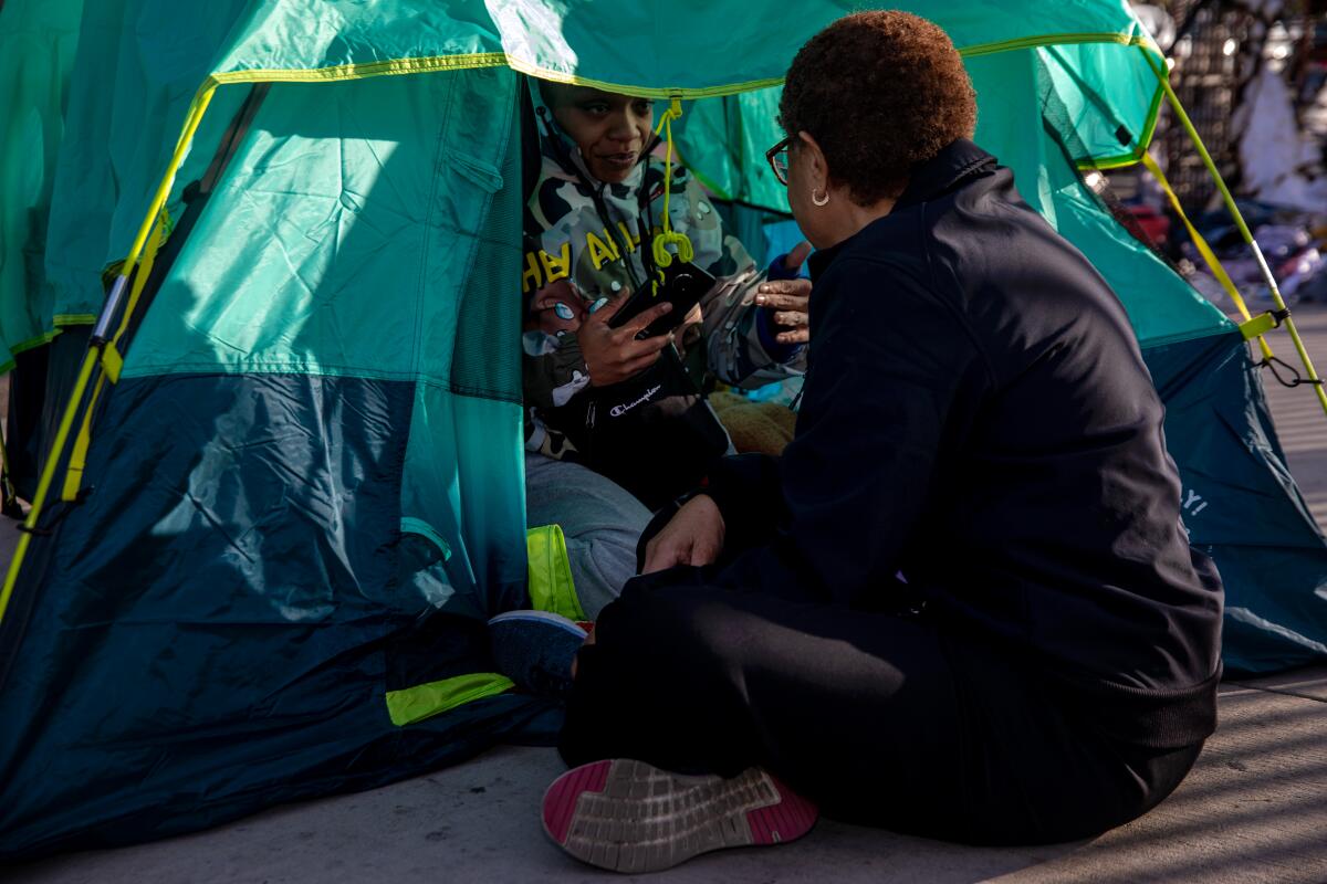 Los Angeles Mayor Karen Bass sitting on a sidewalk by the opening of a tent, chatting with a person inside.