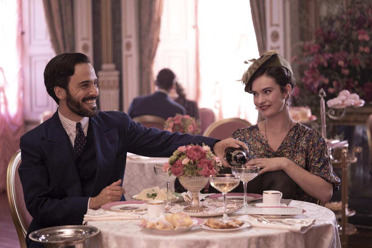 A man pours champagne for a woman in a fancy pink dining room in interwar Britain