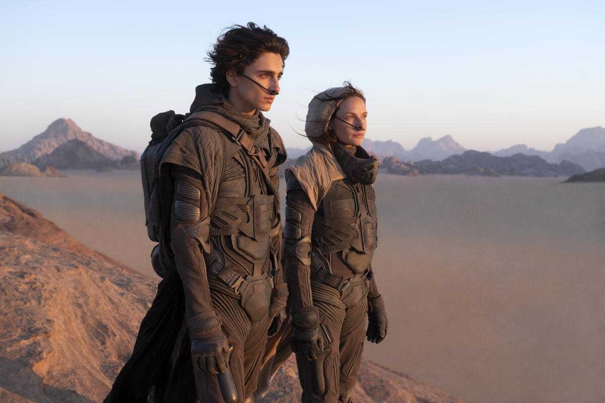 A man and a woman stand on the desert-like surface of an alien planet.