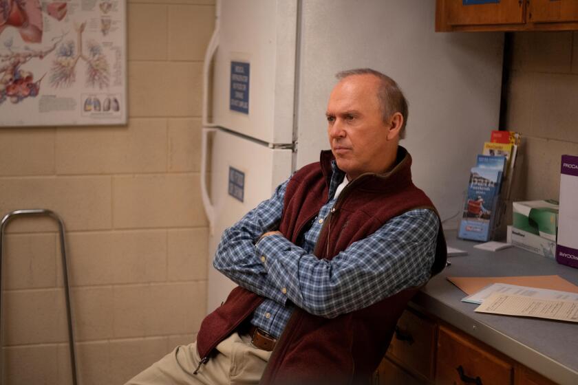 A man in a plaid shirt and fleece vest sits leaning against a counter next to a refrigerator.