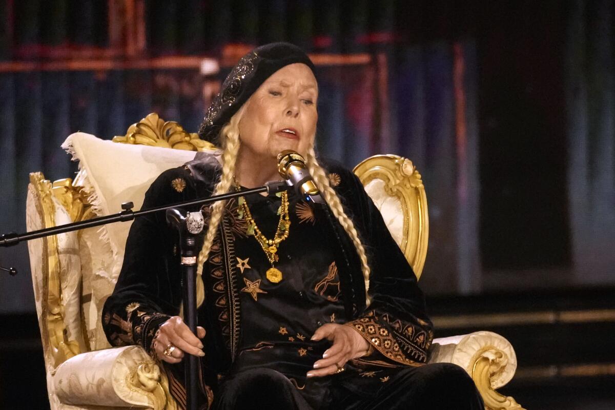 Joni Mitchell in a black dress and hat with long braids sitting in a chair and singing on a stage