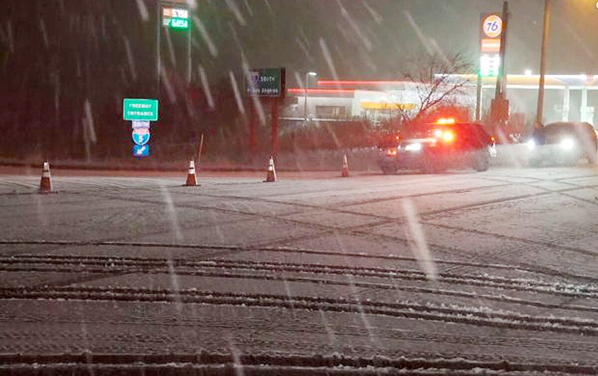 Snow falls on an intersection and freeway onramp closed off by traffic cones and a police cruiser with lights on in the dark