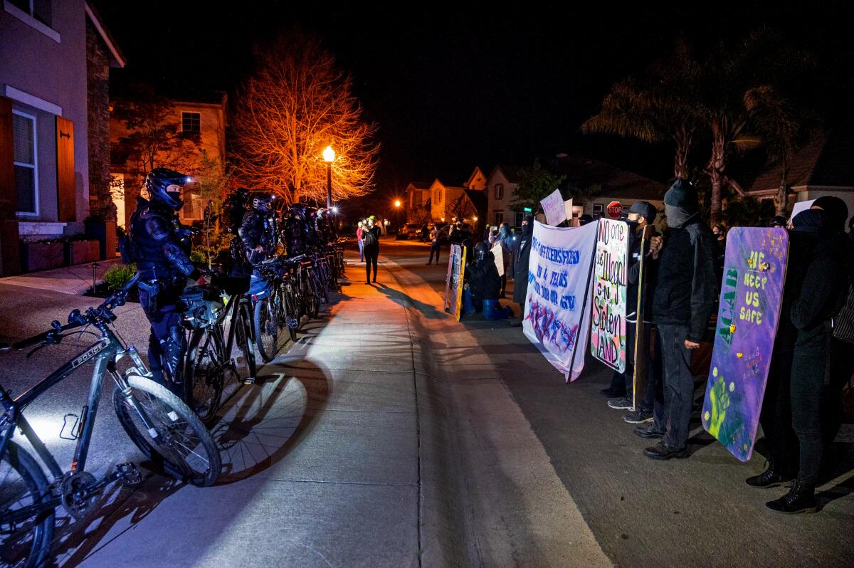 A row of protesters faces off with a row of police officers on a suburban street at night.