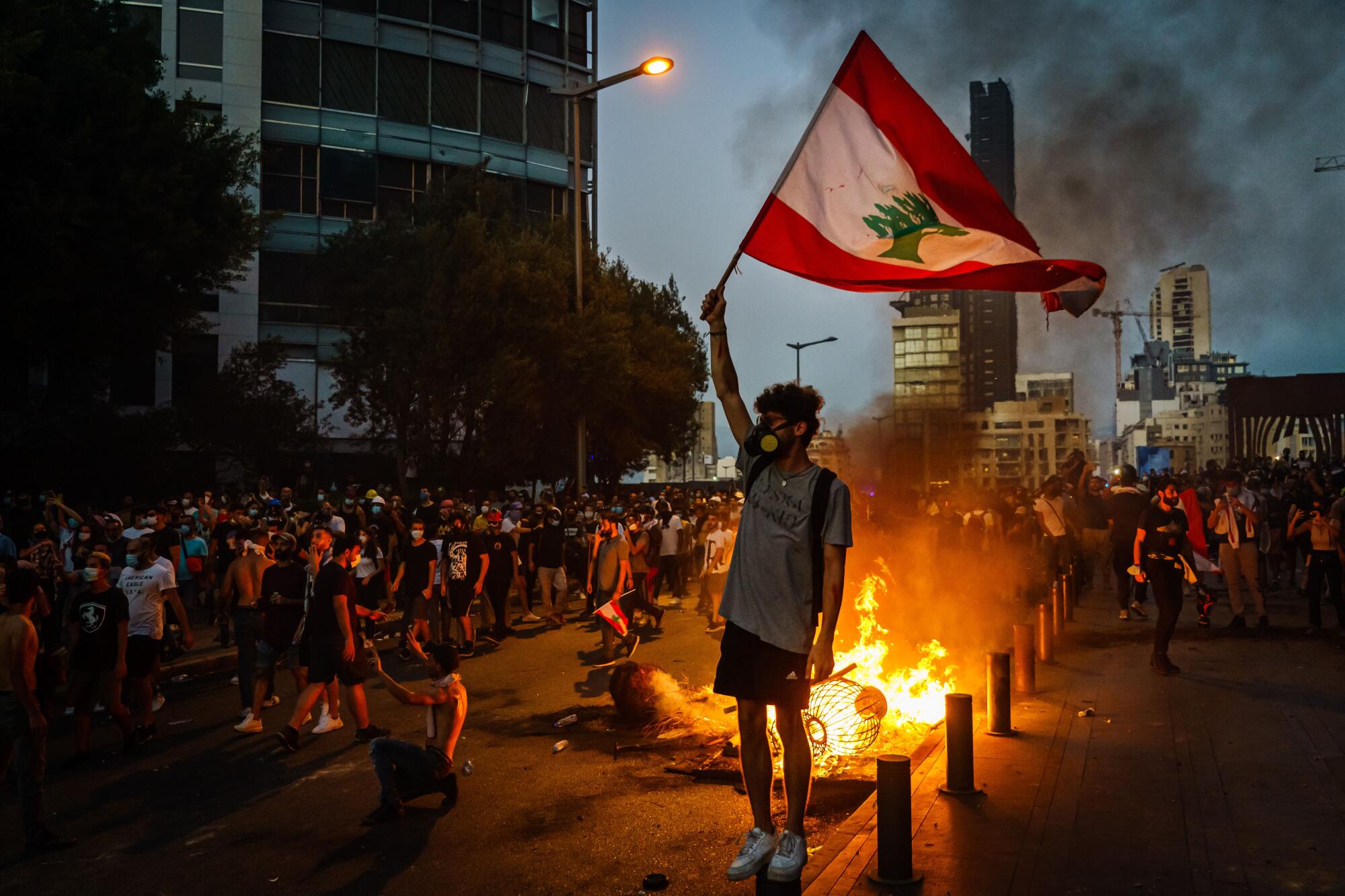 At dusk, a fire glows behind a man in a mask holding a flag high over his head.