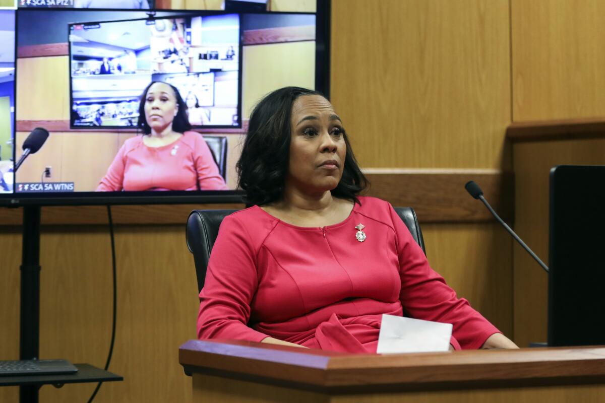 A woman in a red top takes the stand during a hearing. A video screen of her is in the background.