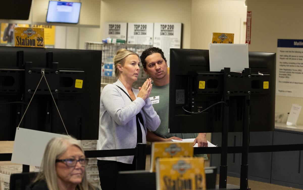 two people stand between two tv monitors in an office setting.