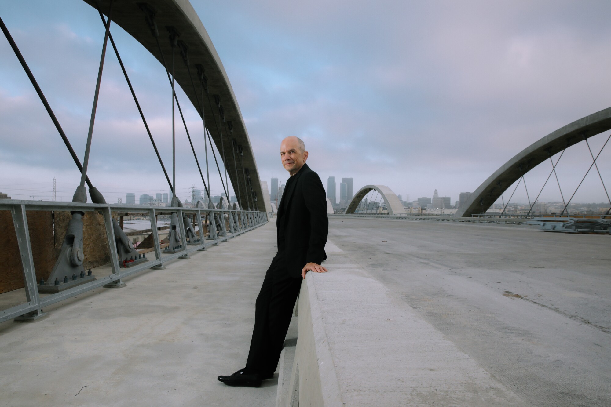 Michael Maltzan, in a black suit, sits on a bridge guardrail.